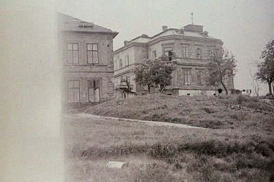 Formal, two-story building with classical detailing & a mansard roof sits atop a small rise. Surrounding foliage obscures por...