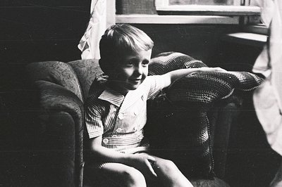 A young boy, likely between 5-8, sits on a plush armchair, smiling slightly. He wears a striped shirt and appears relaxed. Th...