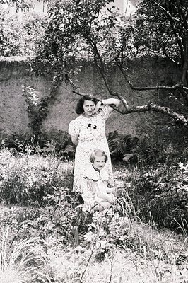 A mother and young daughter in a overgrown garden, the woman with one hand on her hip. Both wear patterned summer dresses. A ...