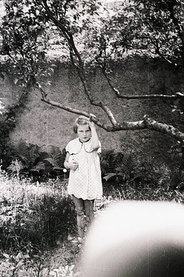 Young girl in a patterned dress stands outdoors, supporting a tree branch with one hand. Lush ferns and foliage fill the back...