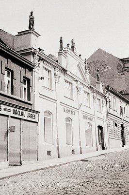 A long view of commercial buildings lining a cobbled street, featuring ornate facades, shop signs ("ÚtČlav JAREŠ," "NÁBYTEK")...