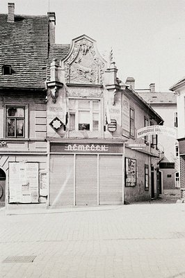 Detailed black and white photo shows a corner building in a European town. Features ornate façade detailing, shuttered storef...