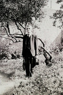 A formally dressed gentleman stands in a meadow, partially obscured by twisting branches and foliage. He wears a dark suit, t...