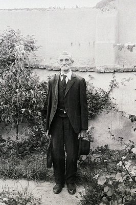 Elderly gentleman in a three-piece suit stands outdoors, holding a briefcase. His posture is formal, suggesting a professiona...