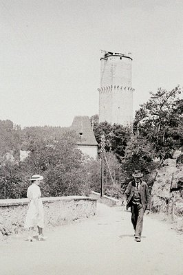 Vintage photograph depicting a stone water tower and surrounding landscape. A woman in a light dress & hat walks away, while ...