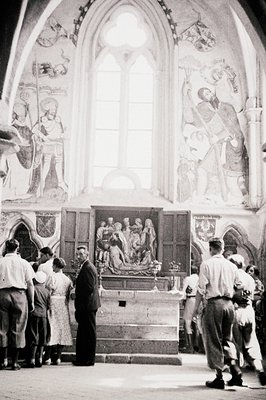 View of an ornate church interior. Figures depicted in murals adorn the high arches. A group of visitors, mostly men in suits...