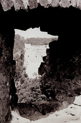 Framed view through a rocky crevice reveals a tiered dam structure and forested landscape. Likely a promotional photo showcas...