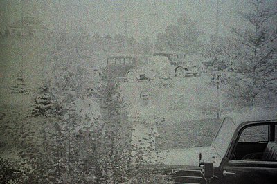 A snow-covered street scene featuring a classic car, likely a sedan, viewed through the windshield. Dense foliage lines the r...
