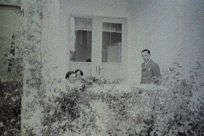 A black & white photo depicts a man in a suit standing near a large window, with a child visible within the room. Exterior wa...