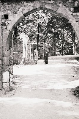 A formal portrait through an ornate stone archway. A man in a suit and hat, leaning on a cane, stands with a woman in a long ...
