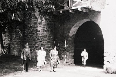 Three figures stand before a large stone archway, seemingly a gate or entrance. The elder man uses a cane, while the women we...