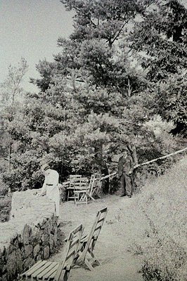 A man in a suit and hat stands near a stone structure and tiered garden beds. Two folding chairs are visible, seemingly aband...