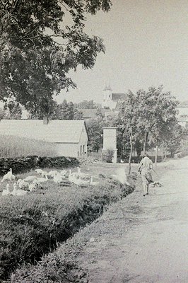 Rural scene: a farmer walks along a dirt road past a barn and grazing sheep. A church steeple rises in the background. Possib...
