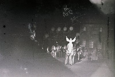 A group gathers outdoors, centered around a man in a theatrical costume with raised arms. Foliage and a building’s façade for...