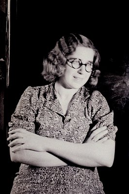 A young woman with a patterned dress and glasses stands with arms crossed, posed indoors. The style of dress and hair suggest...