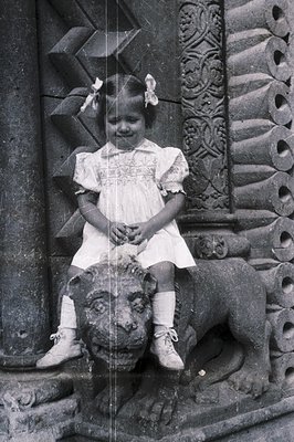 A young girl in a detailed, ruffled dress and white lace-up shoes sits atop a carved stone lion sculpture. The architectural ...