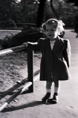 A young child, likely a girl, is captured mid-stride, leaning on a blurred wooden railing. She wears a dark, belted coat and ...