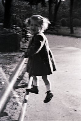A young girl in a dark coat and boots runs past a blurred playground structure. Motion blur suggests rapid movement and captu...