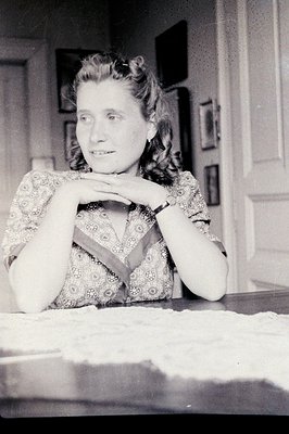 A young woman with styled hair sits behind a table, hands clasped. She wears a patterned dress with short sleeves. Visible in...