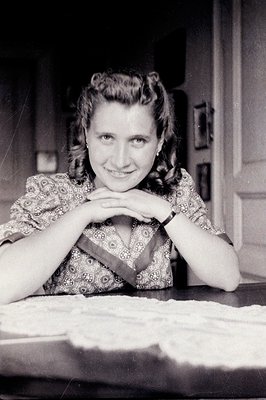 A poised young woman with stylized, curled hair smiles directly at the camera. She wears a patterned blouse and rests her han...