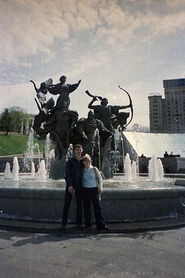 A man and woman stand before a large, allegorical fountain sculpture with tiered water jets. Likely 1970s, this scene appears...