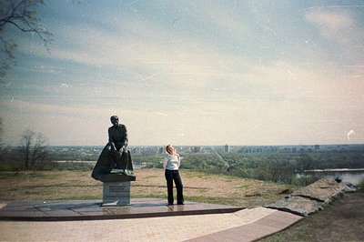 A woman stands posed by a bronze statue overlooking a city panorama. A river bisects the cityscape, marked by buildings and g...