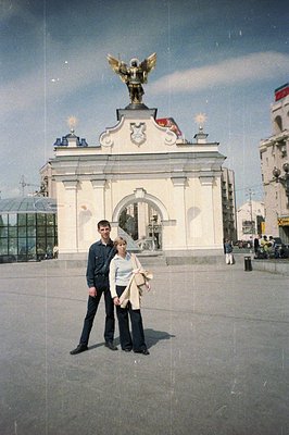 A couple stands before a monumental, neoclassical archway featuring a golden winged statue. The architecture hints at Soviet-...