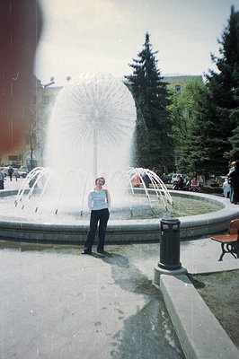 A young woman stands before a large circular fountain featuring multiple jets of water. The scene includes mature evergreen t...