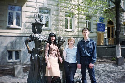 A bronze sculpture depicts a woman in early 20th-century attire, flanked by two young adults in casual 1990s clothing. Buildi...