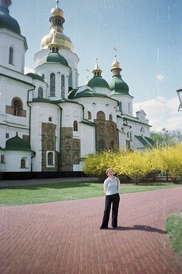 A woman stands before a striking Orthodox church with multiple green and gold domes. The architecture features white walls co...