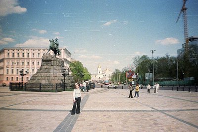 A wide, paved square features a bronze equestrian statue atop a large, tiered base. A woman stands in the foreground. In the ...