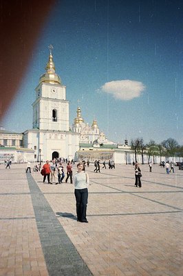 Kyiv Pechersk Lavra monastery complex, featuring a white bell tower & golden-domed churches. A lone woman stands in the foreg...