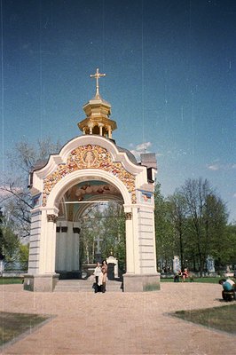 Ornate, whitewashed chapel with gold dome & detailed bas-relief carvings. Two figures stand within the open doorway. Lush gre...