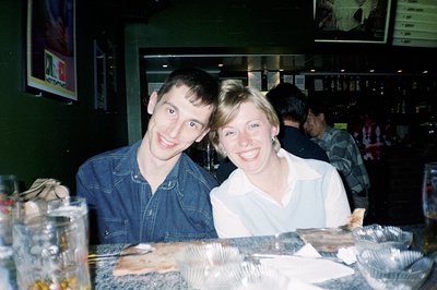 Young man & woman pose at a bar counter; he wears a denim shirt, she a white blouse. Buffet-style trays with food visible. Be...