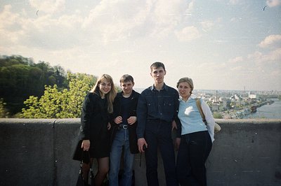 Four young adults pose on a concrete overlook, a river and cityscape sprawling behind them. Denim jackets & dark clothing sug...
