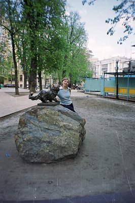 A young woman perches atop a large boulder featuring a bronze statue of a crouching tiger. Lush greenery and a classical buil...