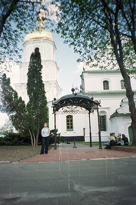View of a classicist-style building with a tall bell tower, possibly a church. A wrought-iron pergola stands in the foregroun...