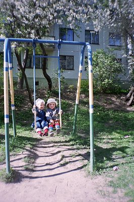 Two young children sit on a simple, metal swing set, likely in a courtyard or park. The background shows a multi-story buildi...