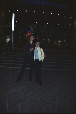 A young couple stands on tiered concrete steps during a rainfall, likely outside a theater or public building. The man wears ...