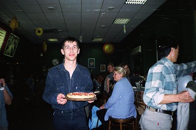 A young man in denim overalls holds a pizza on a tray, presenting directly to the camera. The scene appears to be a casual, d...
