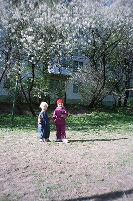 Two young children in coordinating outfits stand in a grassy yard, framed by blooming fruit trees. A mid-century apartment bu...