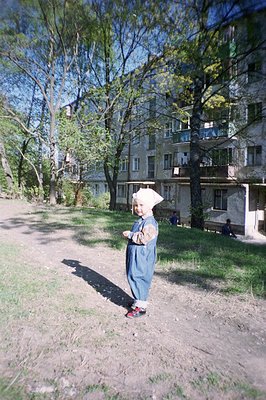 A young child stands in a dirt patch, wearing denim overalls and a patterned shirt, appearing to pose. Lush greenery and a mu...