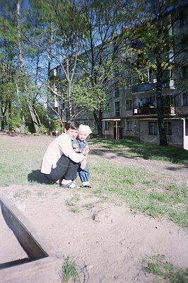 A woman in a light-colored jacket kneels, holding a young boy in denim overalls. They are positioned in a grassy courtyard be...