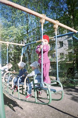 A playground scene with children on a monkey bar structure. A girl in a pink suit & red hat hangs from the top, while three o...