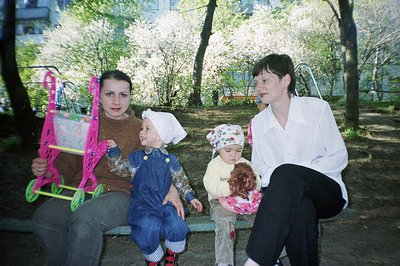 Two women and two young children sit on a park bench beneath blooming trees. One child wears overalls; the other holds a doll...