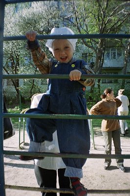 A young child, in a sailor-style dress and dark tights, playfully poses behind a metal barrier. The scene appears to be an ou...