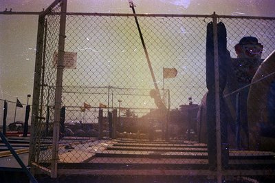 Vintage color photograph captures a deserted amusement park scene, framed by a chain-link fence and a clown cutout. Faded fla...