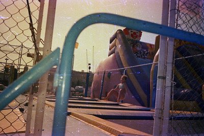 A young woman sits on a staircase near a vibrant, cartoon-themed water slide. Visible behind the chain-link fence, a coastlin...