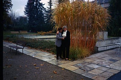 Two women in dark coats stand embracing near a tall ornamental grass display in a formal garden. The setting appears to be a ...