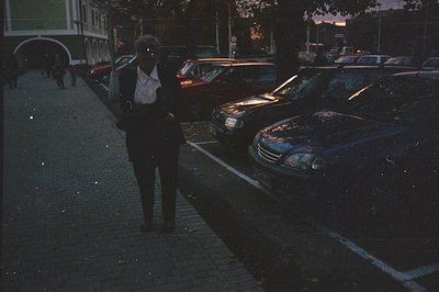 A man in a dark suit and white shirt walks along a wet, cobbled street lined with parked cars. Rain glistens on the vehicles,...